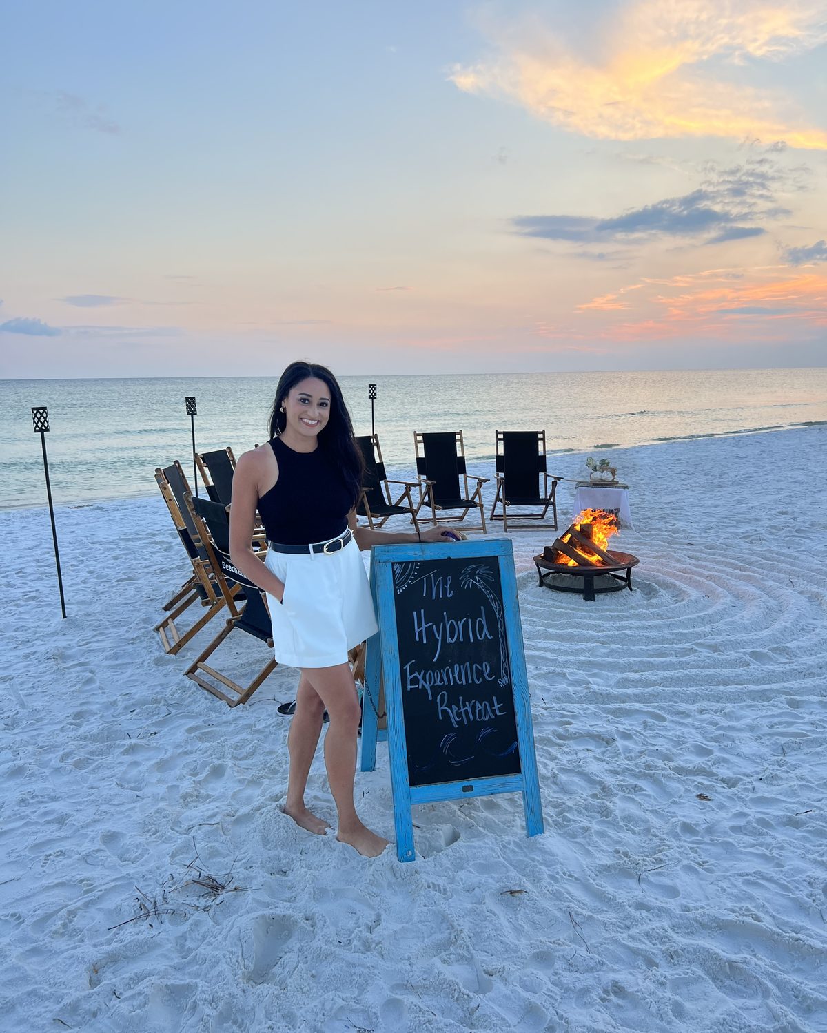Macie beside The Hybrid Experience Retreat sign at sunset, with a beach bonfire and chairs set out behind her